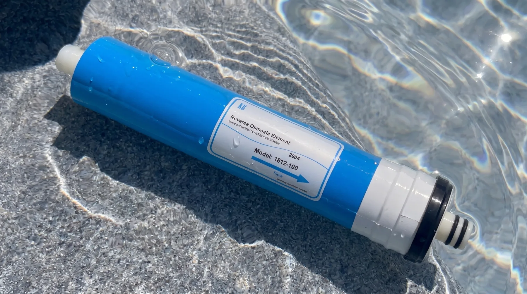A blue reverse-osmosis element lying in shallow sunlit water, its label reading Model 1812-100.